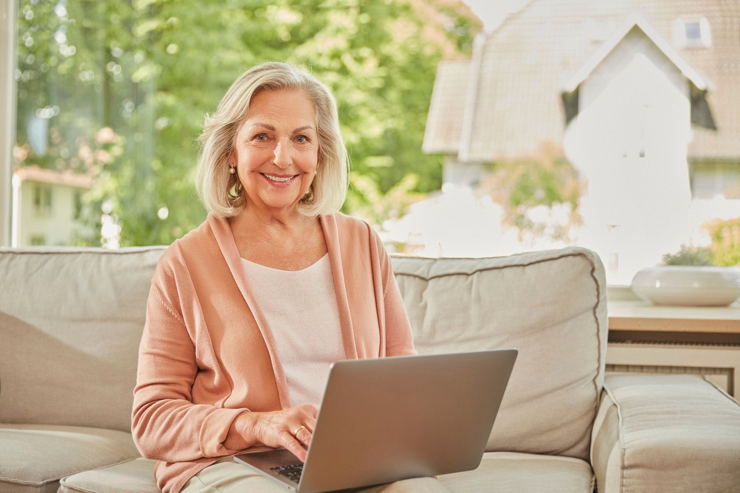Eine Frau sitzt auf einem Sofa am Laptop und lächelt in die Kamera.